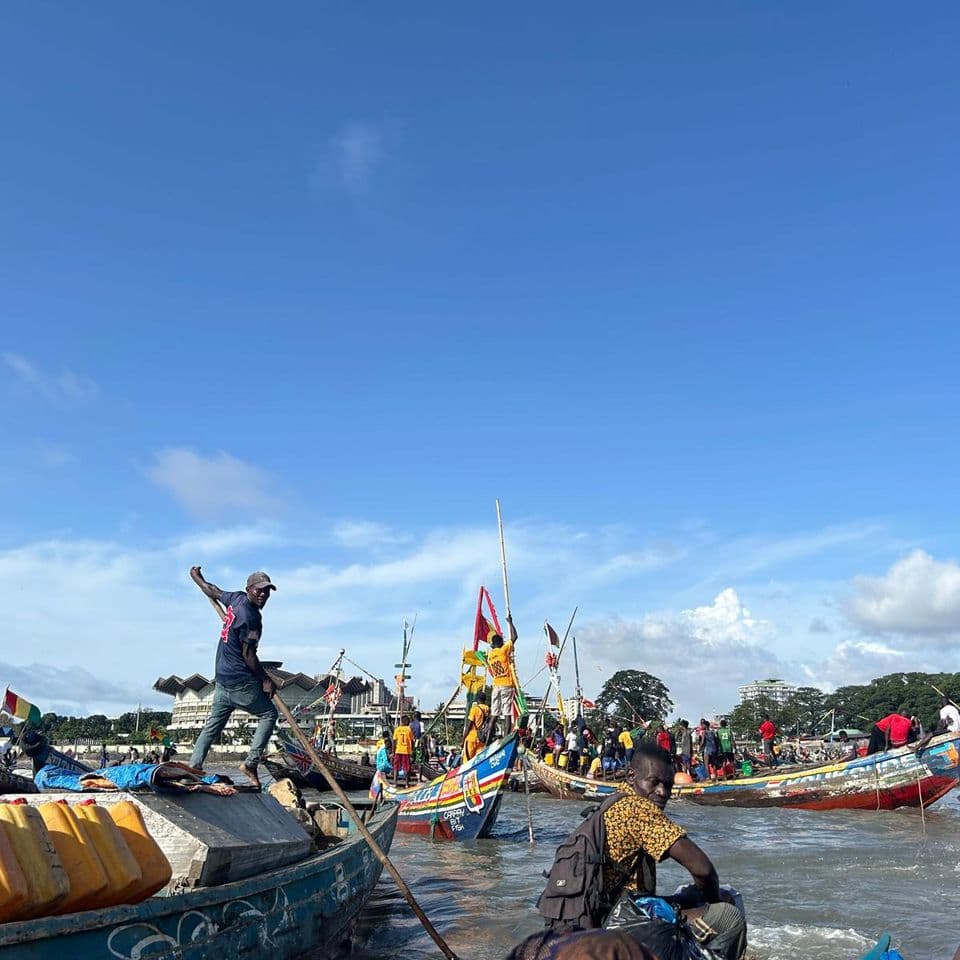 Fishermen on colorful boats at a busy shoreline, with clear blue skies and distant trees in the background.